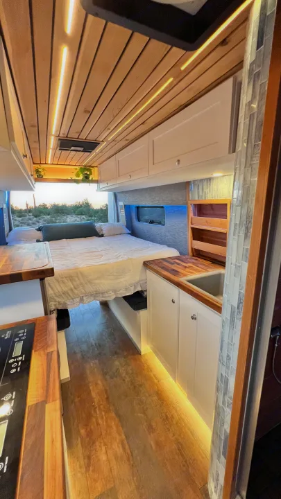 Spacious RV rental interior with a cozy bed, equipped kitchen, wooden ceiling, and warm under-cabinet lighting.. Ford Transit 2019