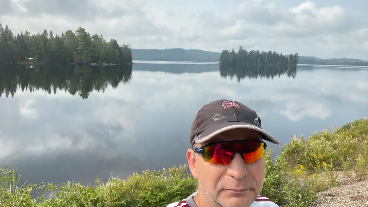 Peaceful lake with mirrored reflections of trees and clouds, seen by a man in sunglasses. An ideal RV rental getaway into nature.. Gulf Stream Grand River 22Mb 2024
