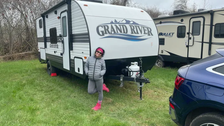A person in a gray jacket and pink boots stands by a white Grand River travel trailer. This RV rental is set up on grass with another RV and a car nearby.. Gulf Stream Grand River 22Mb 2024