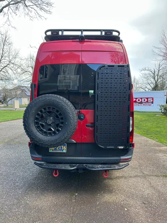 Rear view of a red custom camper van, equipped for travel with a spare tire, gear panel, and roof rack. Perfect for an RV rental adventure.. Mercedes Sprinter 2022