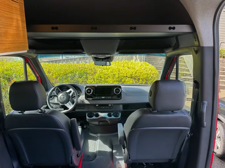Front cabin of an RV rental, showing the modern dashboard, Mercedes steering wheel, and comfortable black seats.. Mercedes Sprinter 2022