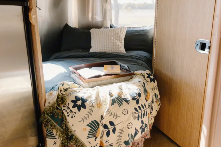 Discover comfort in this RV rental bedroom: dark bedding, floral blanket, and a tray with books, bathed in natural light.. Airstream Flying Cloud 2017