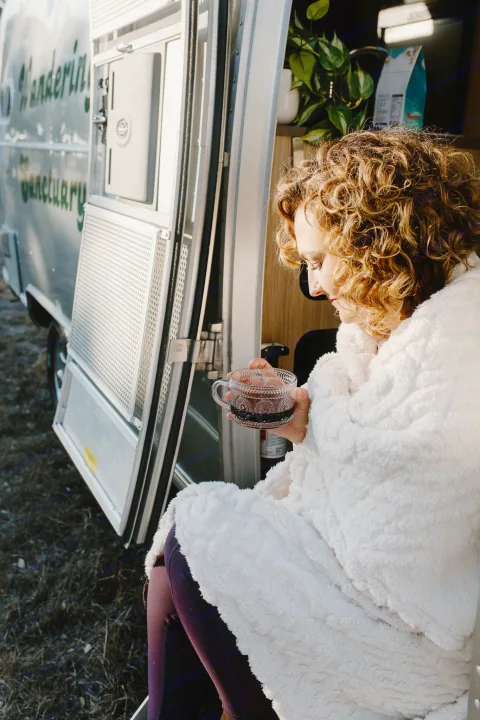 Find your cozy haven with an rv rental! A woman in a fluffy white blanket sips from a mug, sitting in her camper's open doorway.. Airstream Flying Cloud 2017