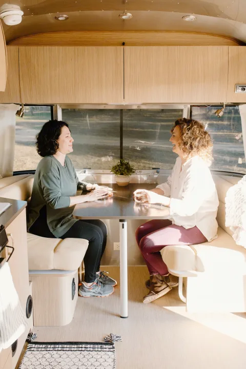 Two women chat inside a bright RV rental with modern wood interior. Coffee & smiles set for a cozy trip.. Airstream Flying Cloud 2017