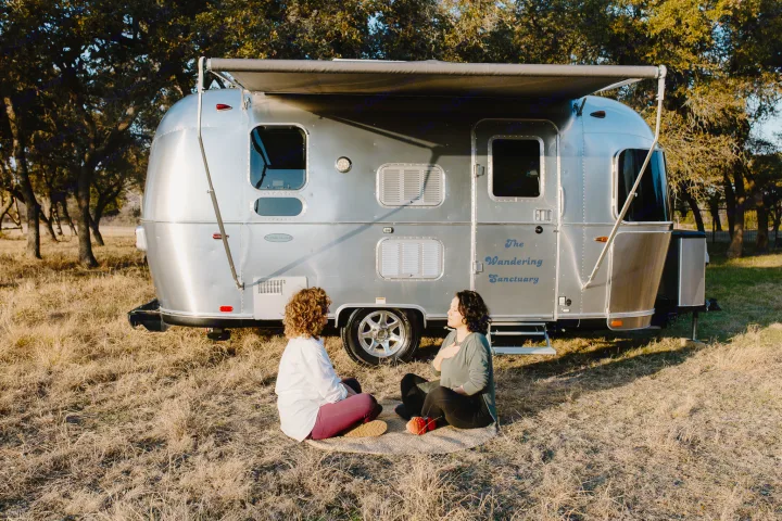 Two women relax by an Airstream RV rental in a natural setting. "The Wandering Sanctuary" is visible on the side.. Airstream Flying Cloud 2017