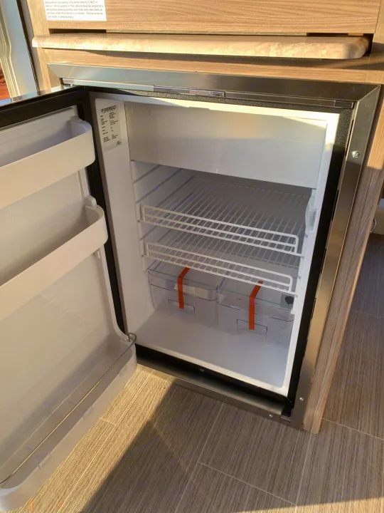 Empty Vitrifrigo refrigerator inside an RV rental, showing wire shelves and orange-taped bins ready for use.. Thor Motor Coach Scope 2023