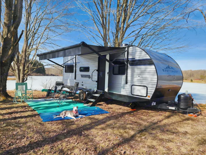This RV rental is set up near a frozen lake, featuring an extended awning, camping chairs, and a dog on a blue mat.. East to West Silver Lake LE 2024