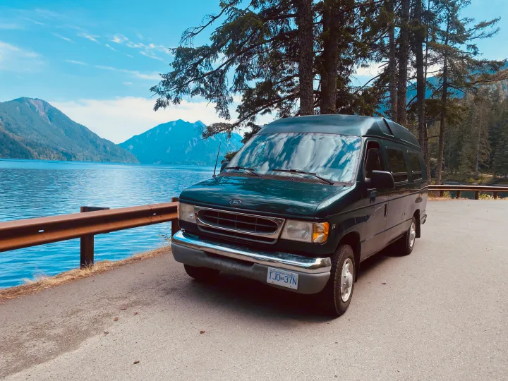 Dark green RV rental van parked by a serene lake, majestic mountains, and lush trees under a blue sky.. Ford Econolline 250s 2001