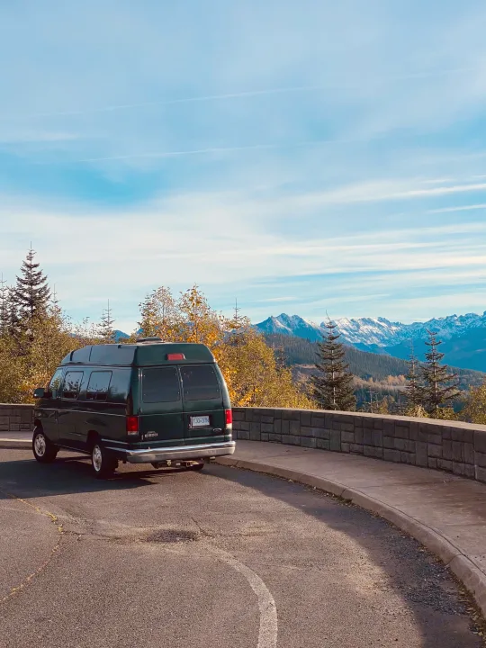 Dark green RV rental van parked at a scenic mountain overlook, with snow-capped peaks and autumn trees under blue skies.. Ford Econolline 250s 2001