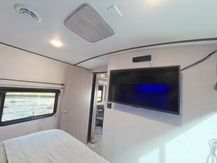 Inside a compact RV rental bedroom, featuring a bed, window with "EXIT" sign, TV, and ceiling AC vent and light, creating a cozy space.. Crossroads Sunset Trail Super Lite 2021