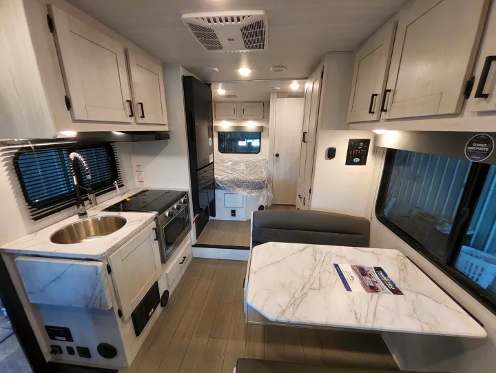 Modern RV rental interior with a compact kitchen, dining booth, and bedroom in the back. Light wood-look floors and white cabinets.. East To West Entrada 2000NS 2025