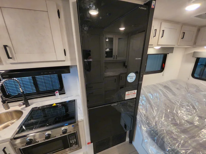 RV rental interior: A compact kitchen with sink, stovetop, and microwave, next to a tall Furrion refrigerator and a covered bed.. East To West Entrada 2000NS 2025
