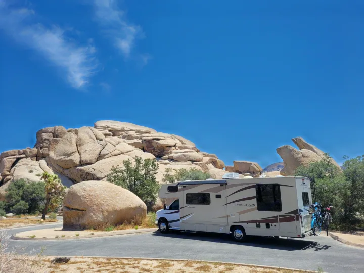 A cream-colored RV rental with bikes parked among massive rock formations & a Joshua tree under a blue sky.. Forest River Coachmen Leprechaun 2018