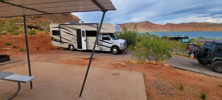 An RV rental is parked at a spacious lakeside campsite, featuring a picnic table, shelter, and scenic mountain views.. Forest River Coachmen Leprechaun 2018