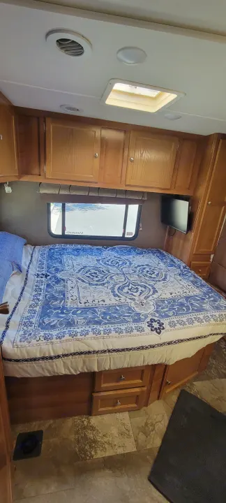 Cozy RV rental bedroom with a large bed, blue patterned comforter, overhead wood cabinets, window, and TV.. Forest River Coachmen Leprechaun 2018