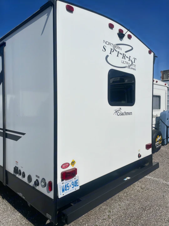 Rear of a white Northern Spirit travel trailer, ready for rv rental. Visible are its window, Ontario plate, and utility hookups.. Coachmen 2557RB 2021