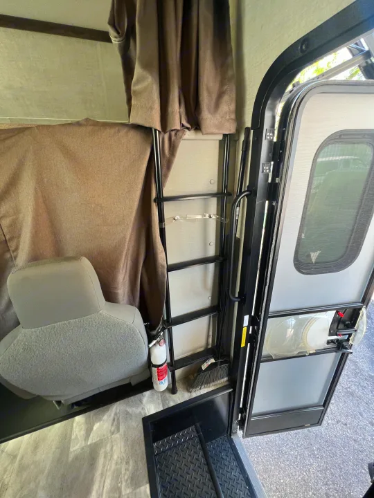RV rental interior: the entrance area with an open door, a ladder, fire extinguisher, broom, and gray seat.. Gulf Stream Conquest 2023