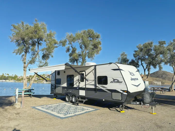 A Jayco travel trailer RV rental parked at a lakefront campsite, awning extended, under a clear blue sky.. Jayco Jay Flight SLX 2022