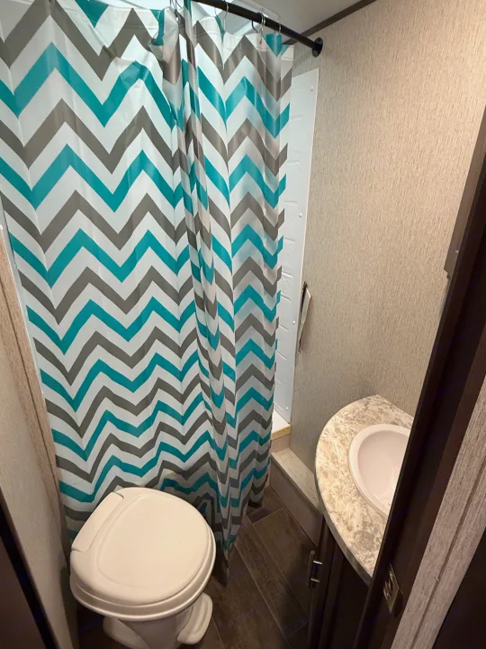 Compact RV rental bathroom showcasing a teal, grey, and white chevron shower curtain, toilet, and sink area. Ideal for travel.. Crossroads Sunset Trail Super Lite 2019