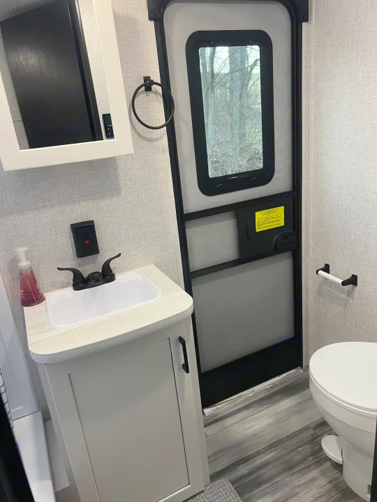 Inside a modern RV rental bathroom: white sink, toilet, vanity, and a door showing a nature view outside.. Jayco Jayflight 324bds 2025