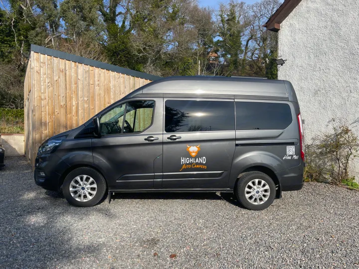 A grey Ford Transit Custom campervan, branded Highland Auto Campers, is parked on a gravel driveway, ready for rv rental.. Ford Transit Custom 2 berth Hi-line Automatic 2021