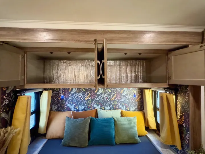 Cozy RV rental interior showcasing a custom floral wallpaper, colorful pillows, and open overhead cabinets with reflective insulation.. Jayco Redhawk 2021