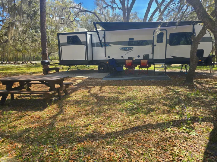 Perfect rv rental setup: White travel trailer with awning, picnic table, & chairs at a shaded, leafy campsite.. Forest River Salem Platinum 2024