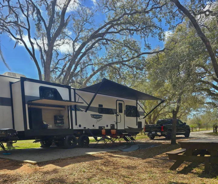 An RV rental, a white Salem Platinum travel trailer, set up at a sunny campground site, surrounded by large, mossy trees.. Forest River Salem Platinum 2024