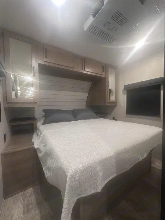 Modern RV rental bedroom with a large bed, light bedding, dark pillows, overhead cabinets, and AC unit. Comfortable travel awaits.. PALOMINO 28BHSS 2024