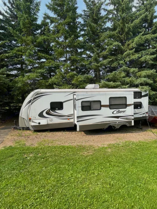 This white travel trailer RV rental is parked with its slide-out extended, nestled by tall trees and green grass.. Keystone RV Cougar 2012