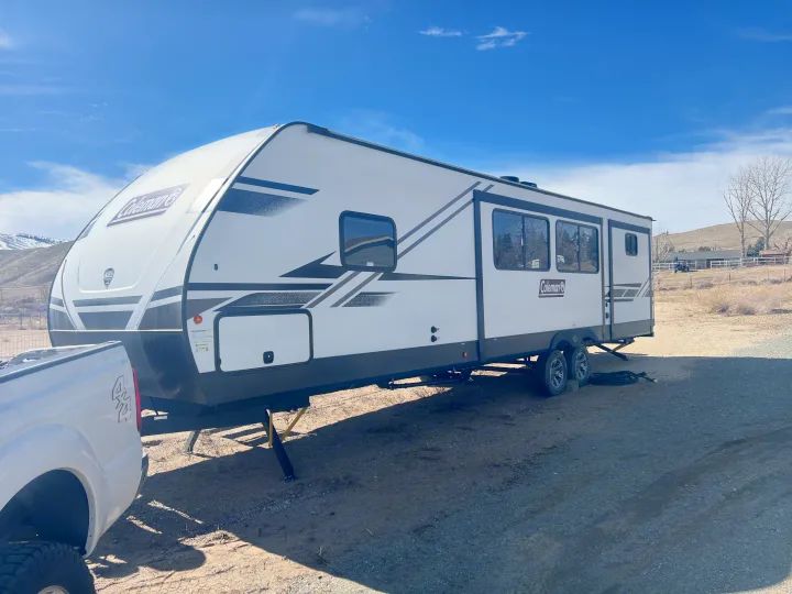 Spacious Coleman travel trailer, ready for an RV rental adventure. Parked on a sunny dirt lot with a white pickup truck; hills in the background.. Coleman 3215BH 2022