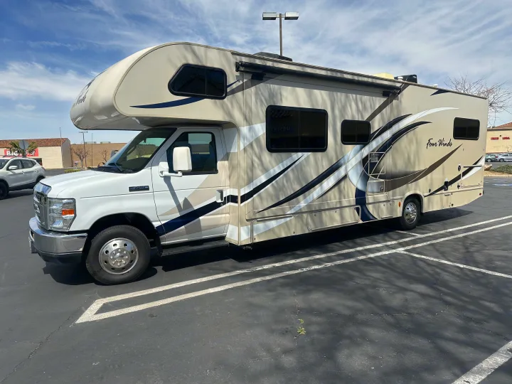A large beige and white Class C RV, perfect for an RV rental, is parked in a lot under a blue sky.. Thor Four Winds 2018