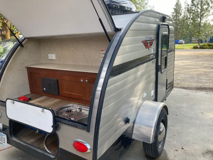The open hatch reveals a compact outdoor kitchen with a sink, stovetop, and storage in this Retro Jr. travel trailer, perfect for an rv rental.. Riverside RV Retro 2017