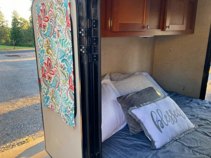 Inside an RV rental: a cozy bed with pillows, a vibrant curtain, and a view of the gravel campsite outside.. Riverside RV Retro 2017