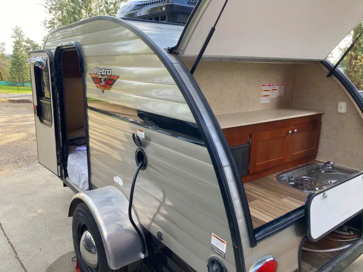 This RV rental teardrop camper features an open side door to a cozy bed and a rear outdoor kitchen with stove and sink.. Riverside RV Retro 2017