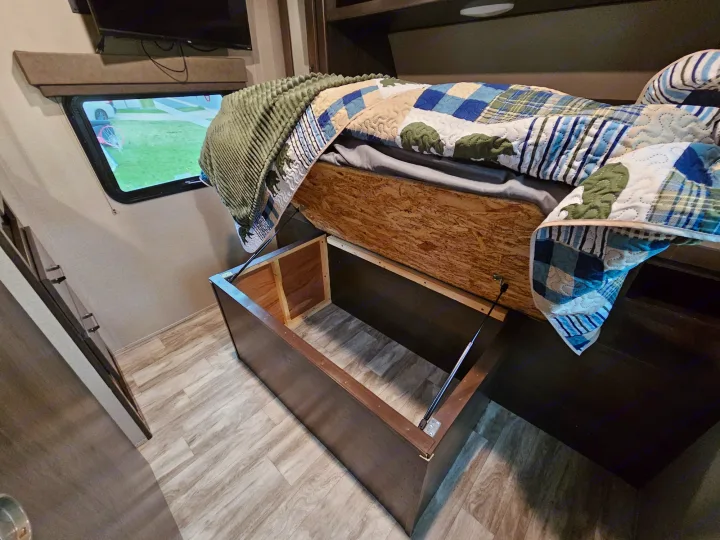 RV rental bed lifts to reveal spacious storage. Features a patterned quilt, window view, and wood-look flooring.. Grand Design Transcend Xplor 2022