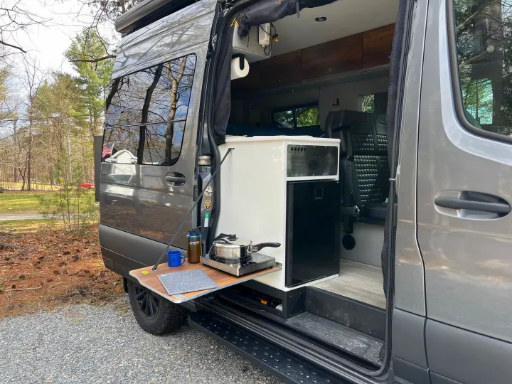 An RV rental camper van showcases its outdoor kitchen with a cooktop on a pull-out table, parked in nature.. Storyteller Overland Stealth Mode 2023