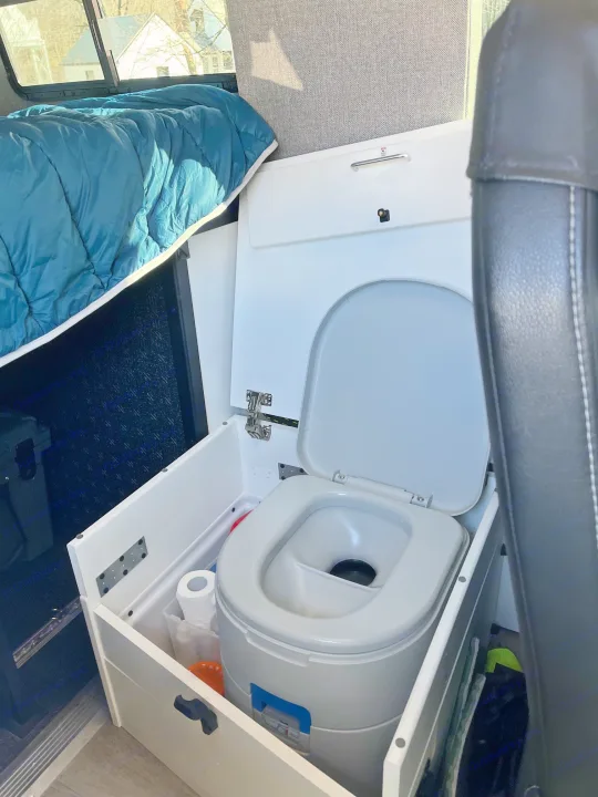 Compact RV rental: an integrated portable toilet with supplies, beside a cozy bed. Ideal for adventures.. Storyteller Overland Stealth Mode 2023