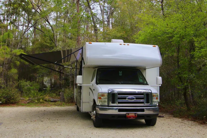 A white Class C rv rental with an extended awning is parked on a gravel lot amidst green trees at a campground.. Forest River Coachmen Freelander 2011