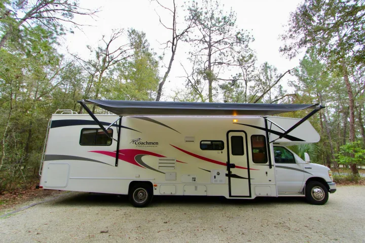 A spacious white RV rental with an extended awning, parked on gravel in a natural, tree-filled camping setting.. Forest River Coachmen Freelander 2011