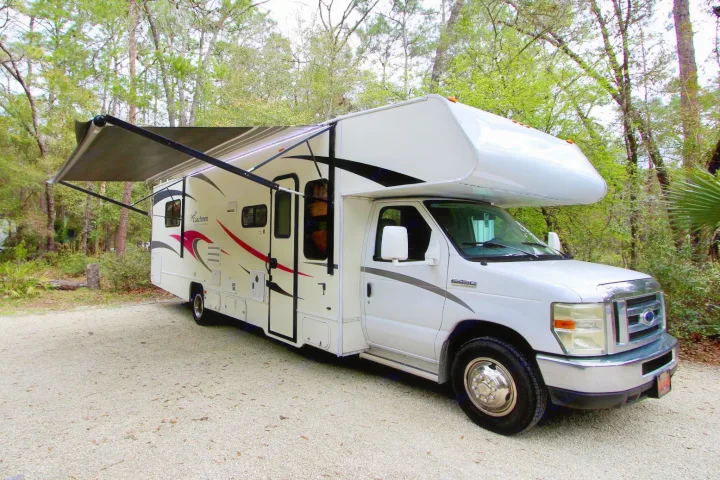 White RV rental with an extended awning parked on gravel amidst trees, perfect for camping and outdoor leisure.. Forest River Coachmen Freelander 2011