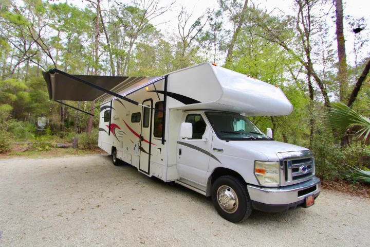 White Class C RV rental parked on a gravel campsite with its awning extended, surrounded by trees, ideal for travel.. Forest River Coachmen Freelander 2011
