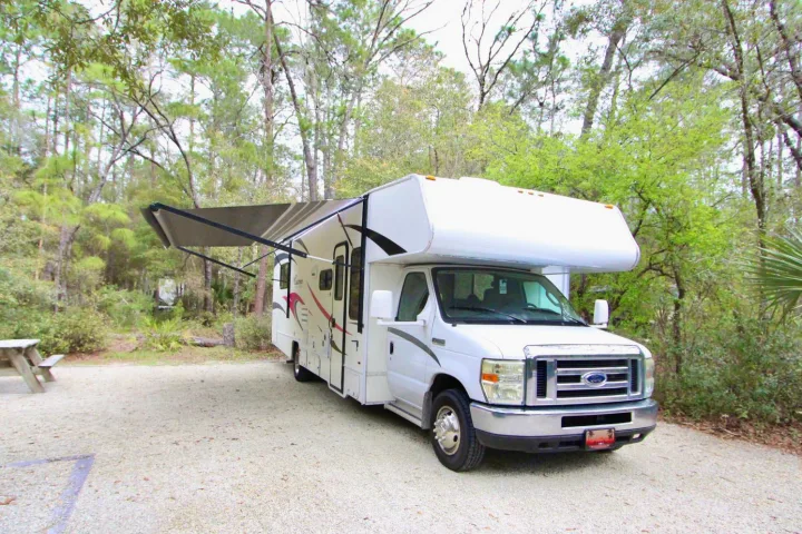 A white Class C RV rental with its awning extended, parked on a gravel campsite surrounded by trees.. Forest River Coachmen Freelander 2011