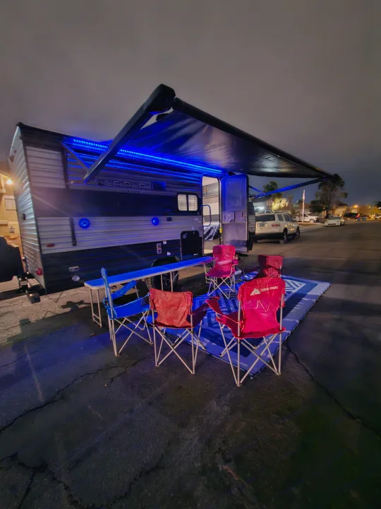 Night view of an RV rental with blue LED awning lights, outdoor table, and red & blue camping chairs set up for a cozy evening.. Forest River Cherokee Wolf Pup 2022