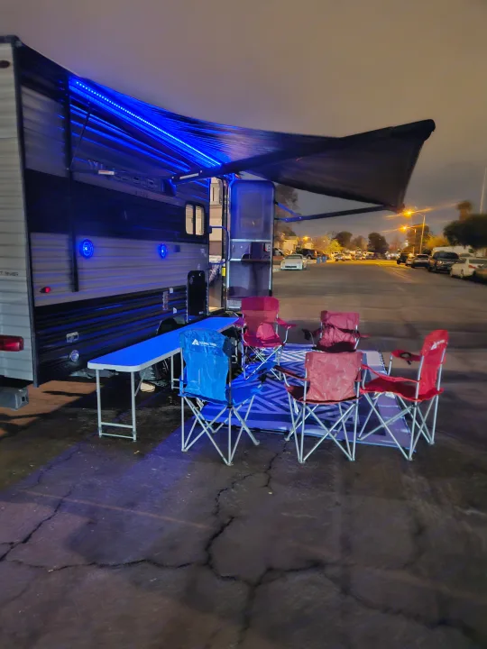 An RV rental campsite at night, featuring blue awning lights, a table, and red & blue camping chairs on a mat.. Forest River Cherokee Wolf Pup 2022