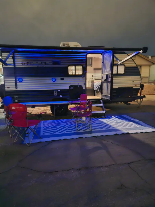 Illuminated RV rental at night with awning, chairs, and an outdoor rug, creating a welcoming camping setup.. Forest River Cherokee Wolf Pup 2022