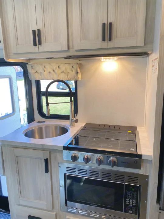 Inside a bright RV rental kitchen: modern sink, Magic Chef stove, Greystone microwave, & light wood cabinets.. Forest River Sunseeker LE 2021