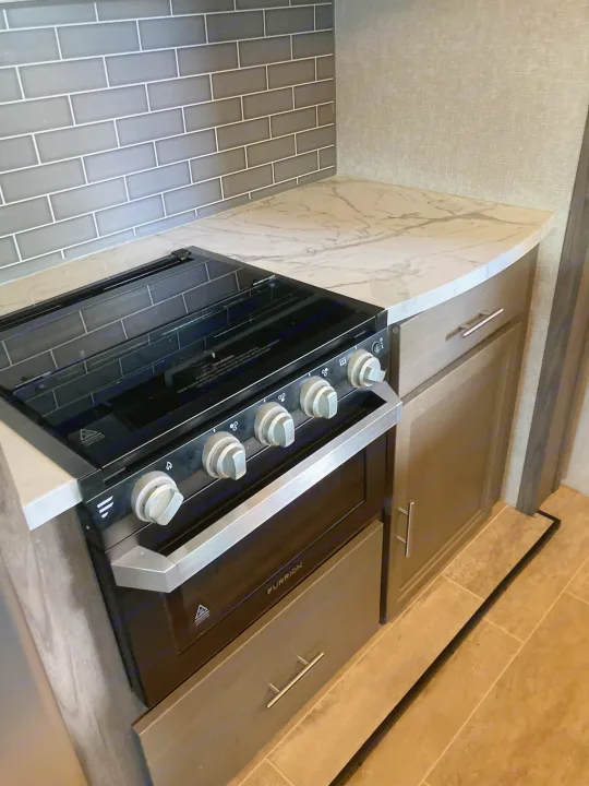 A clean, modern RV rental kitchen featuring a black Furrion stovetop/oven, marble-style countertop, and grey subway tile backsplash.. Dutchmen Kodiak Ultimate 2020