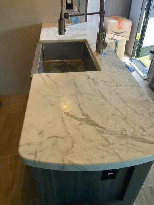 This RV rental kitchen features a bright white countertop with gray veining, a large stainless steel sink, and a modern faucet.. Dutchmen Kodiak Ultimate 2020