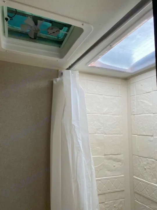 View of an RV rental shower area, featuring a ceiling fan vent, clear skylight, and a white curtain on a curved rod.. Sunset Park RV Sun Lite 2021